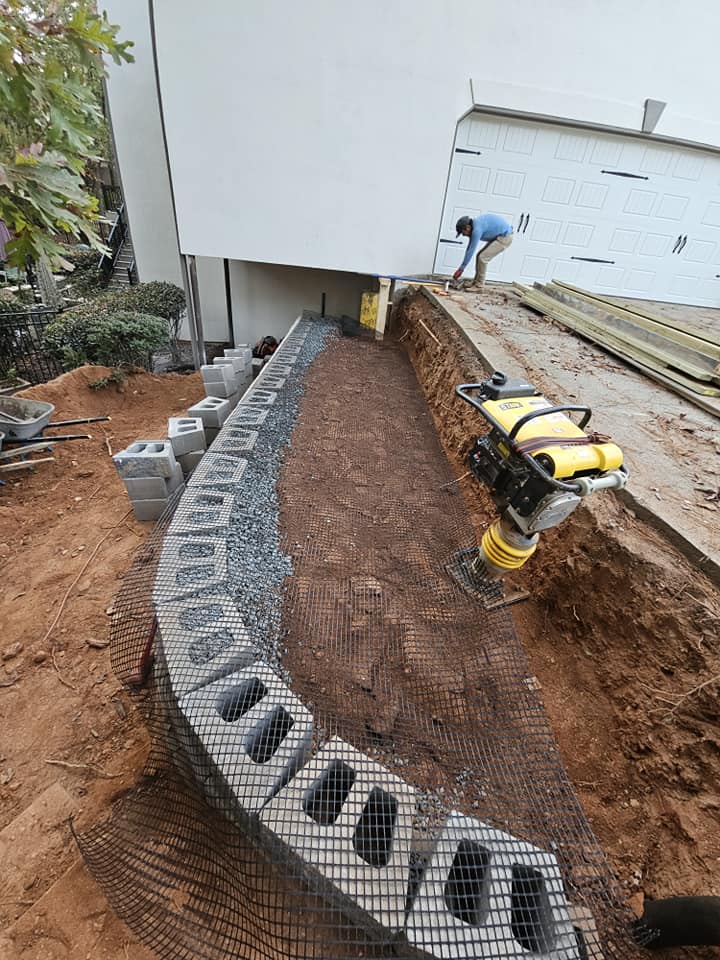 Construction site showing a worker leveling soil and laying concrete blocks for a foundation.
