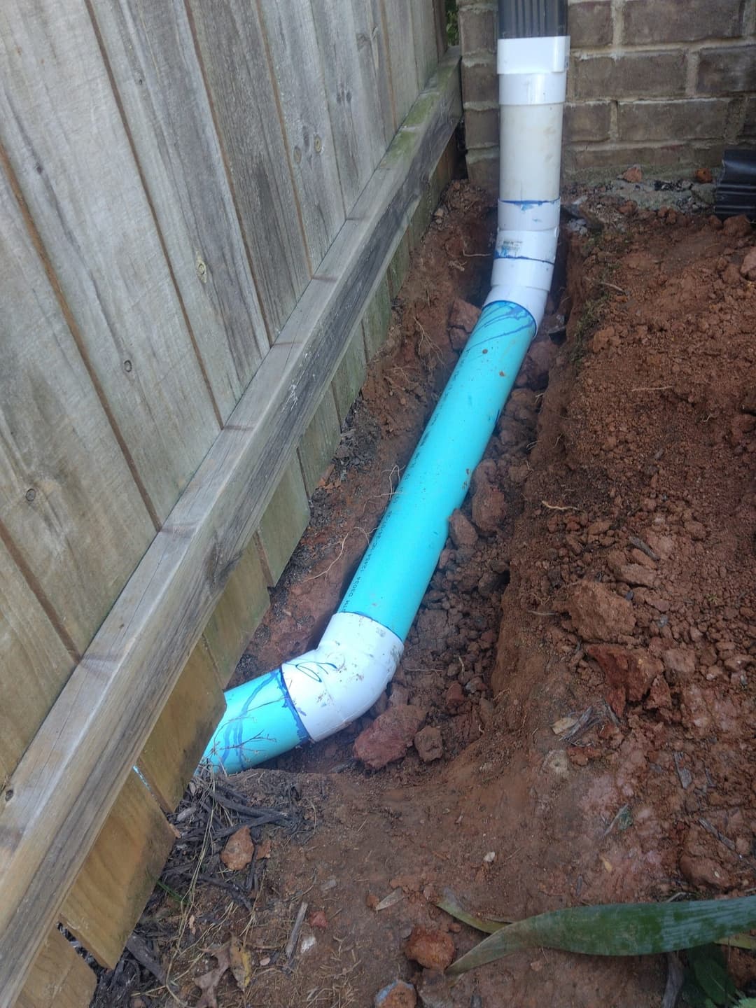 Underground blue PVC pipe installed alongside wooden fence in excavated soil.