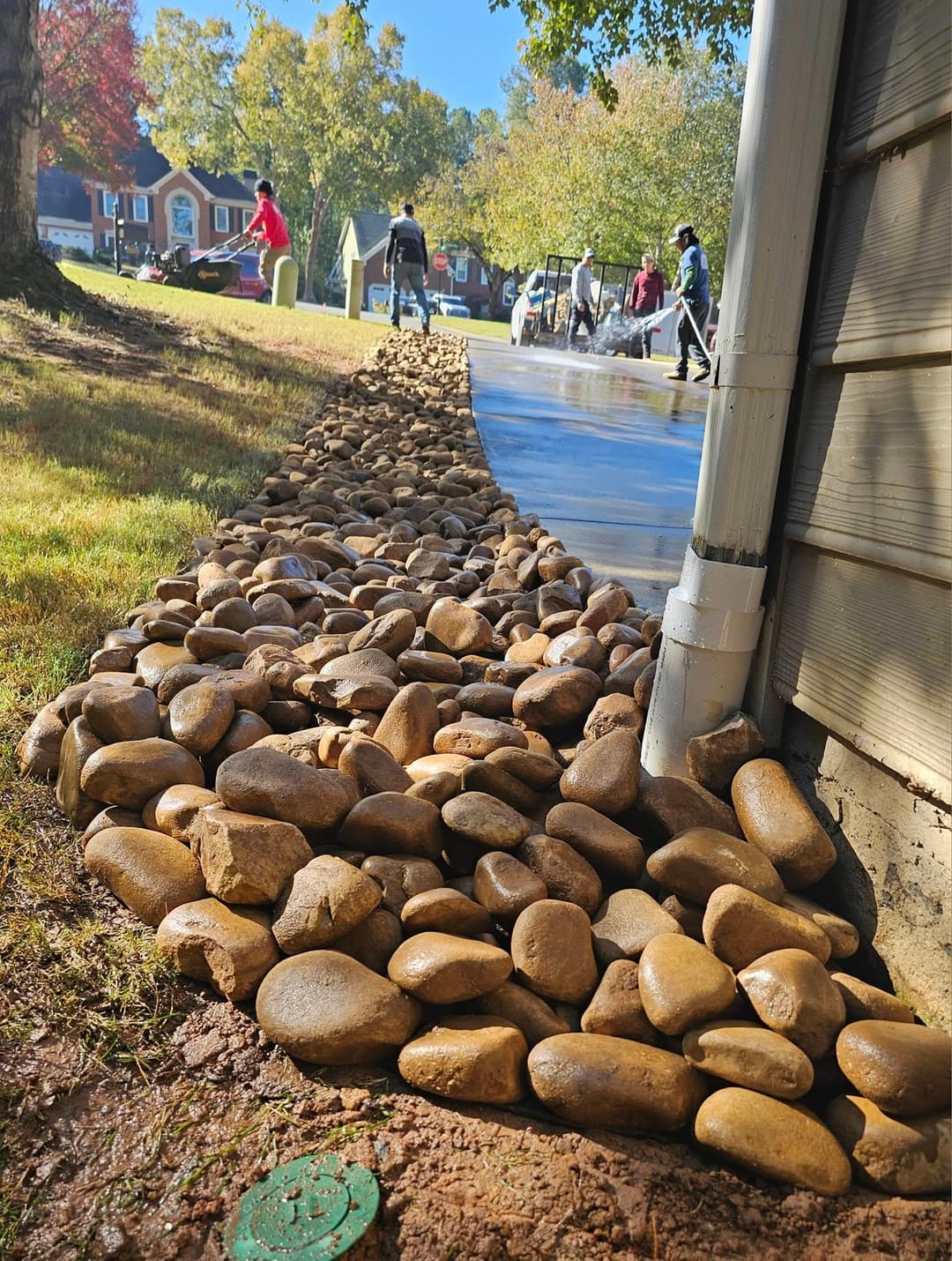 Landscape with a gravel pathway, workers, and a house in a sunny residential area.
