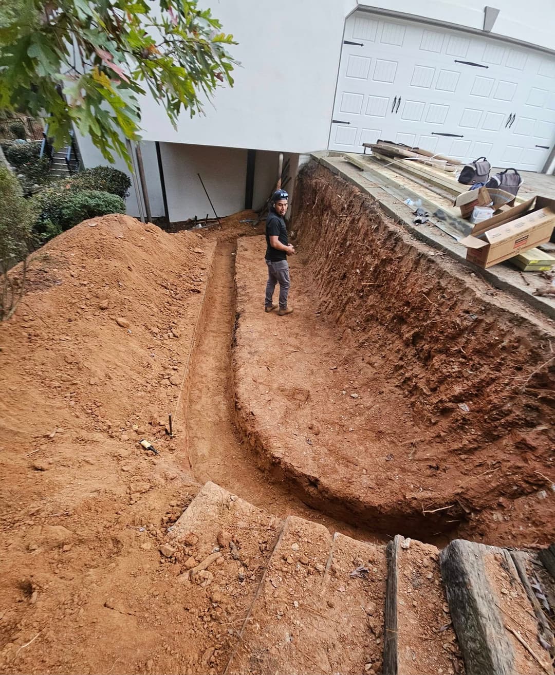 Construction worker standing in a freshly dug trench beside a house, surrounded by dirt and materials.