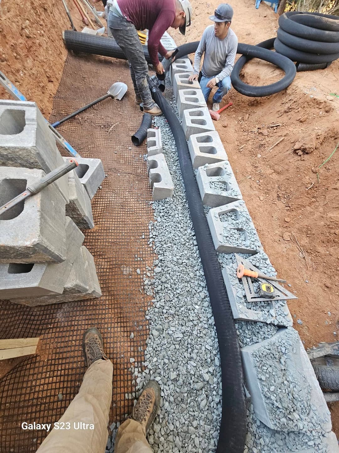 Construction workers installing drainage pipe and blocks on a gravel foundation at a job site.