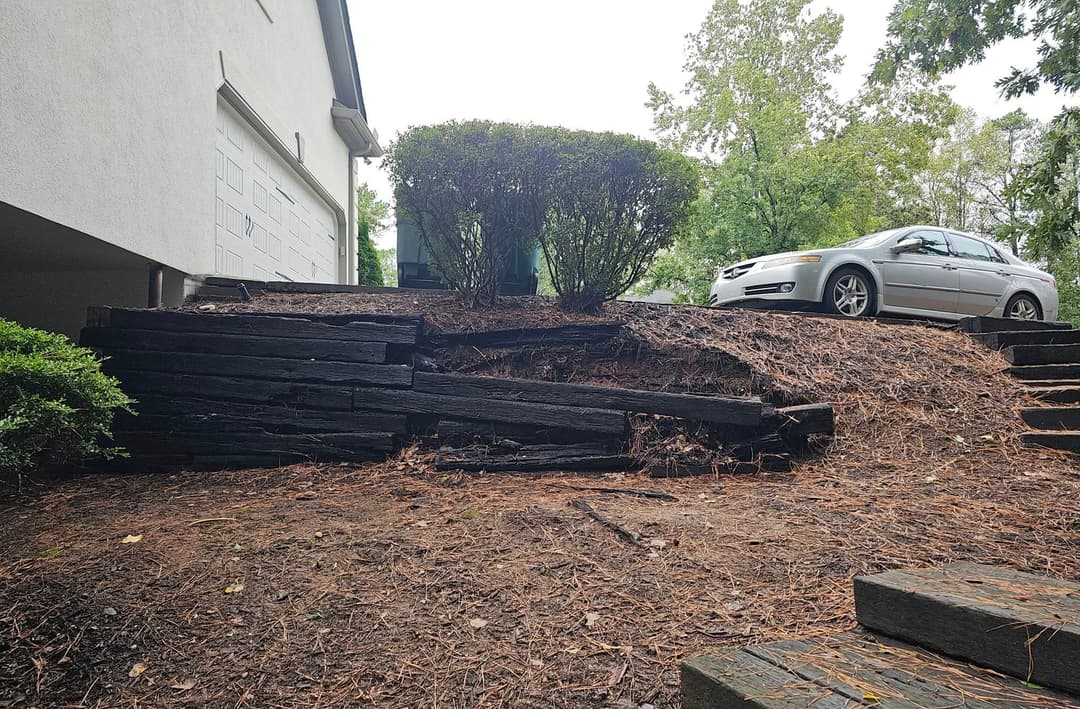 Deteriorating landscaping timbers beside a driveway with a parked car and shrubs.