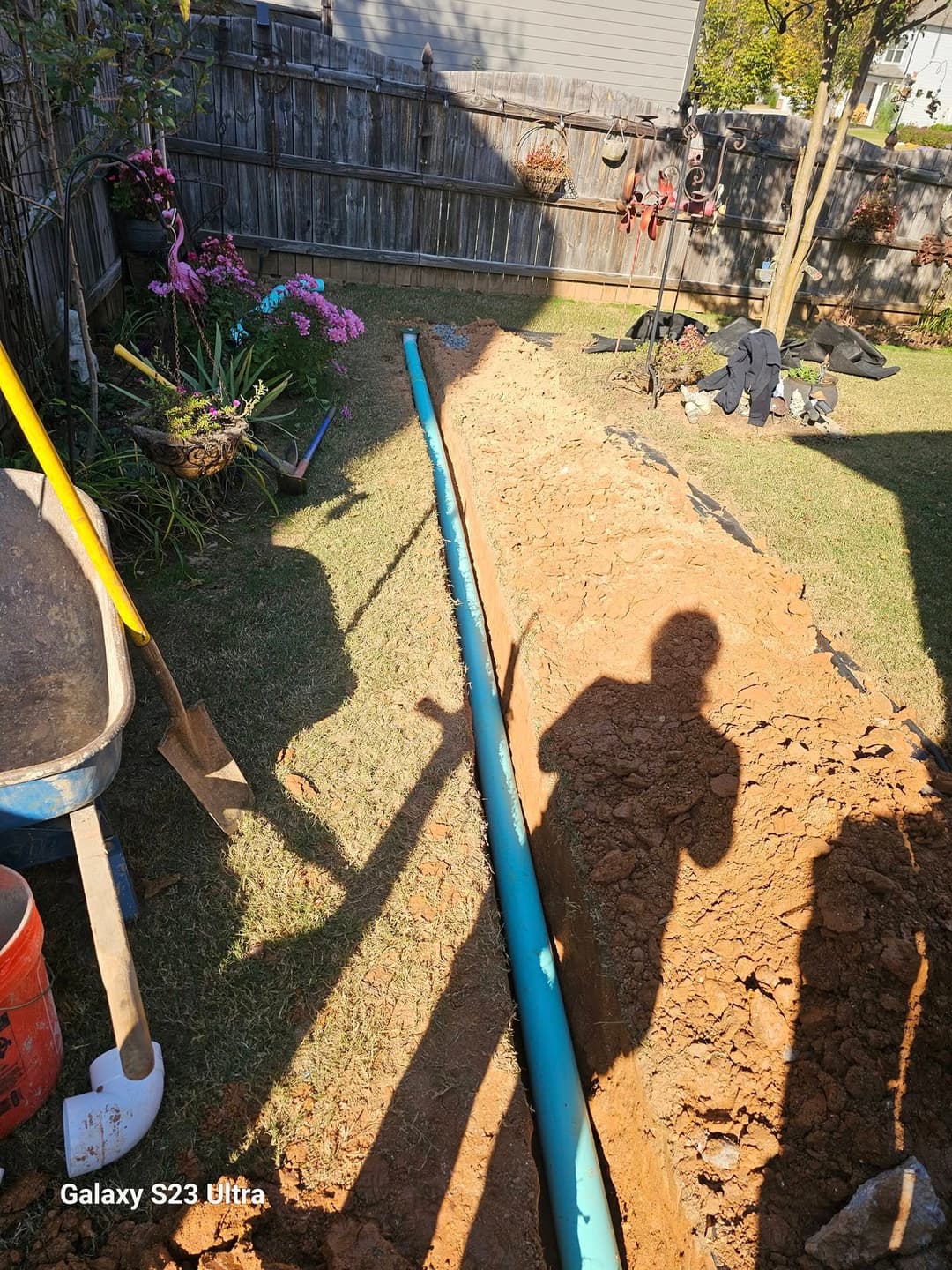 Trenched yard with a blue pipe, tools, and flowers in the background. Ready for plumbing installation.