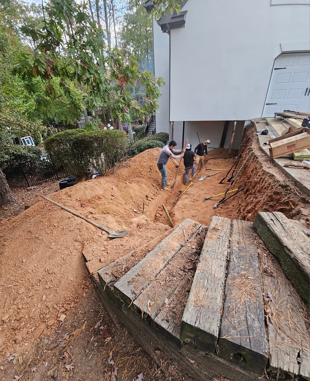 Workers digging a trench near a house foundation, surrounded by trees and wooden planks.