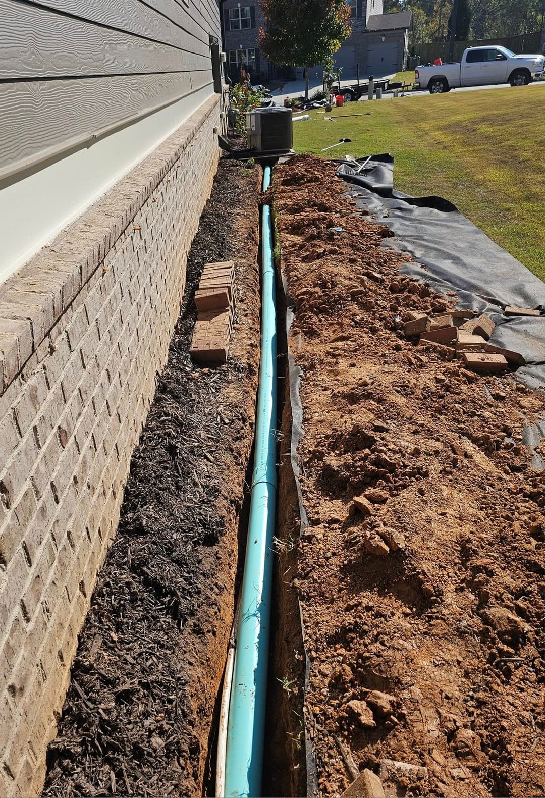 Excavated trench with blue drainage pipe alongside a house, surrounded by soil and landscaping materials.