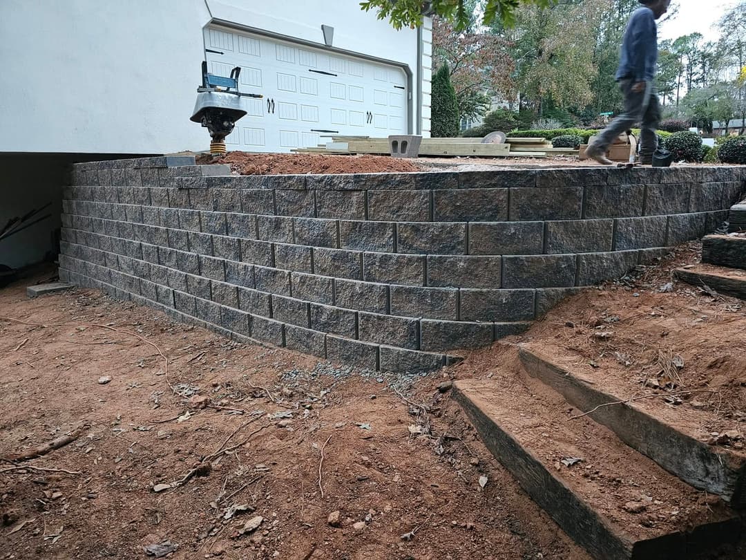Construction of a stone retaining wall with a worker and tools in a residential setting.