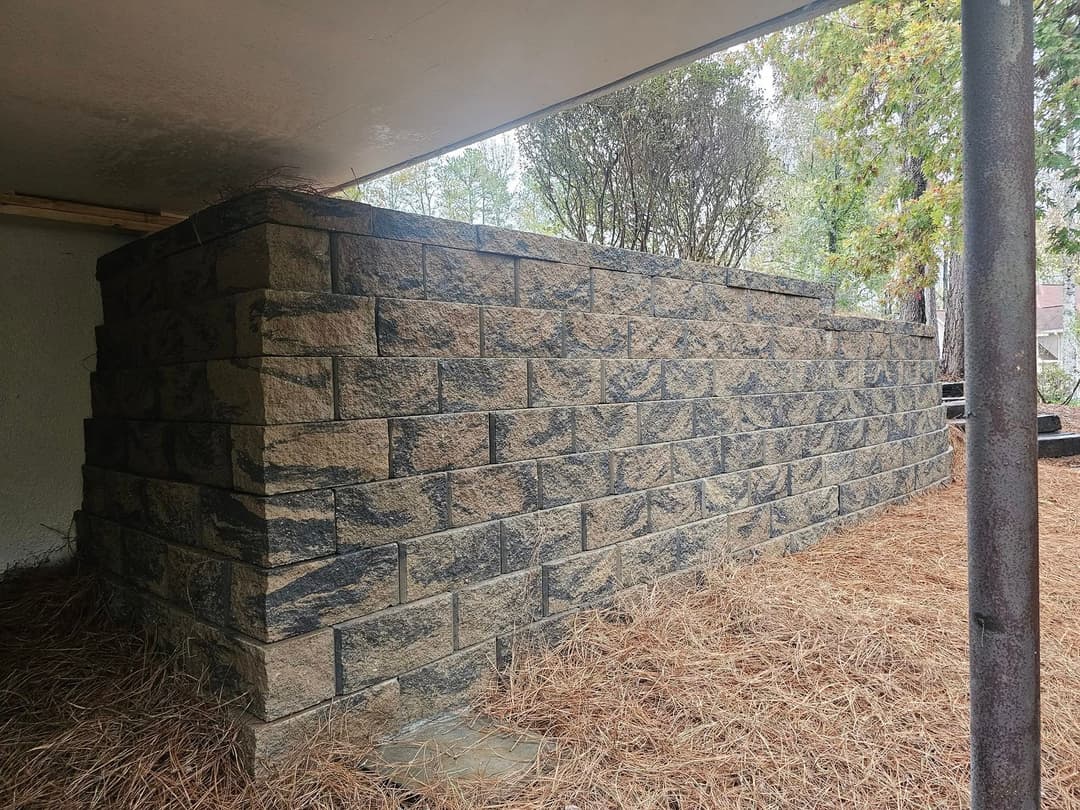 Stone retaining wall surrounded by pine needles and trees under a covered patio area.