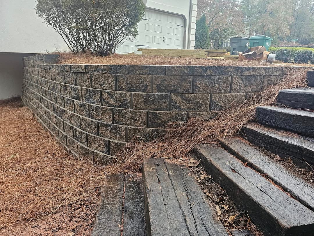 Retaining wall with stone blocks and wooden steps, surrounded by pine needles and shrubbery.