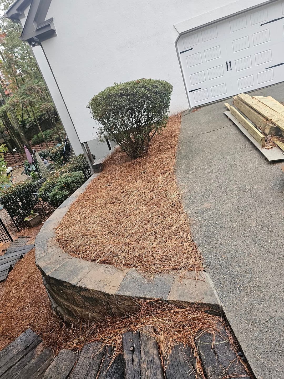Landscape featuring a stone border, pine straw, and a bush beside a driveway and garage.
