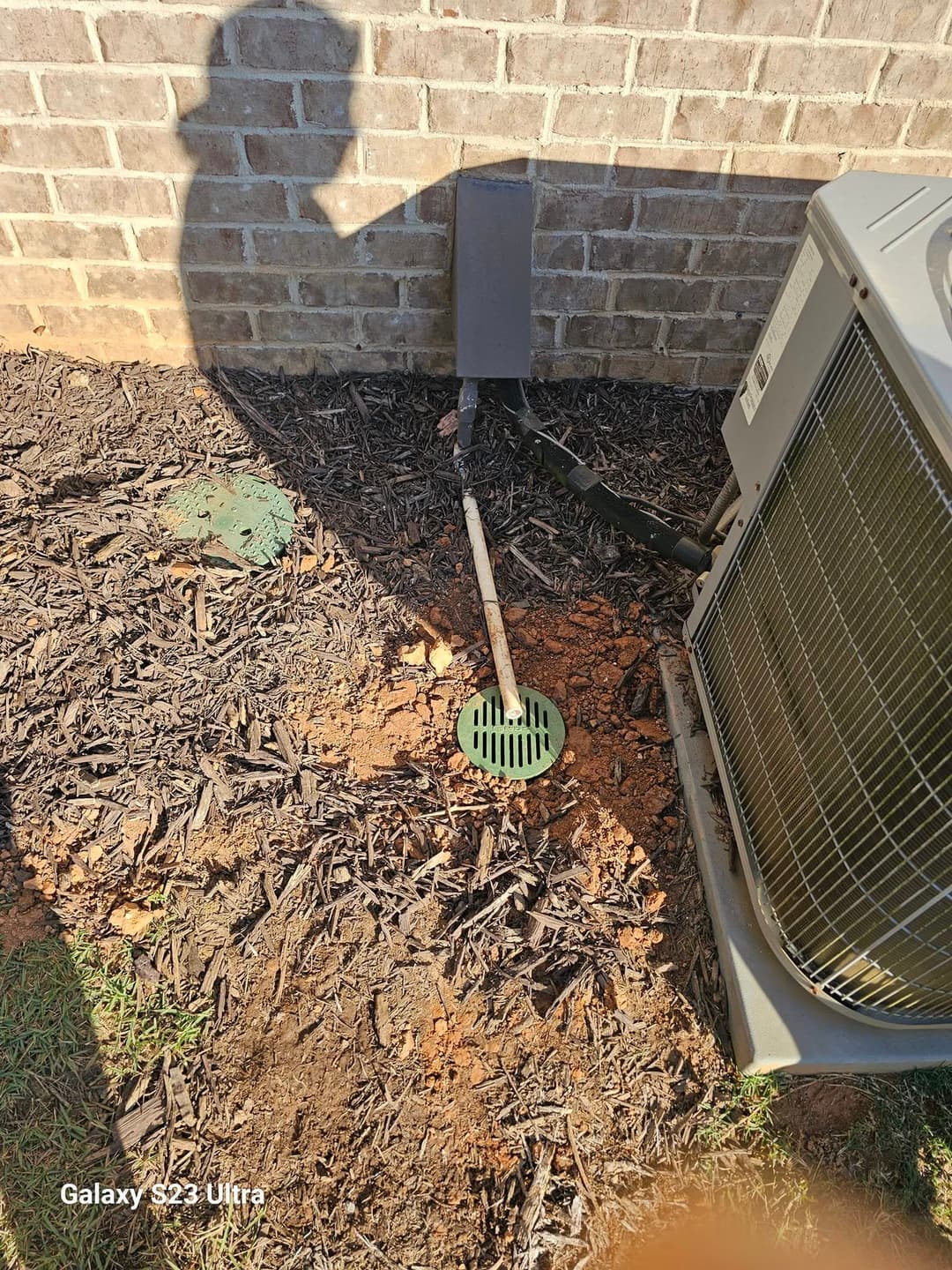 Air conditioning unit near drainage pipe and green grate on mulch landscape.